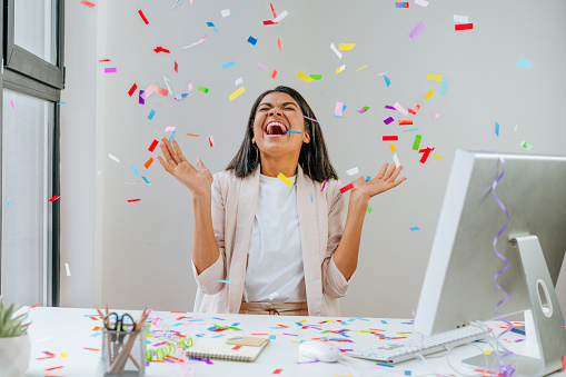 Young business woman having fun time catching confetti sitting at the desk in the office. Party time on the work place. Selective focus.