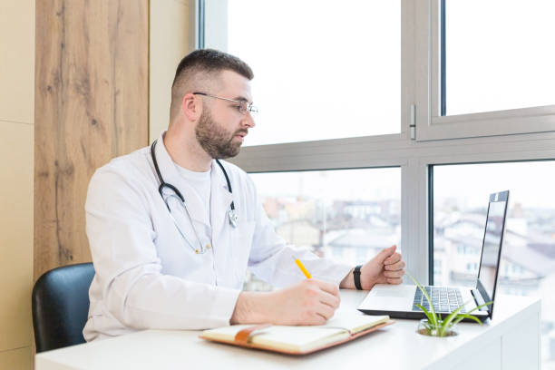 istock Young male doctor working in office on laptop and documents, looking at camera, smiling 1368641400