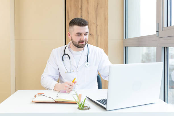 istock Young male doctor working in office on laptop and documents, looking at camera, smiling 1368641403