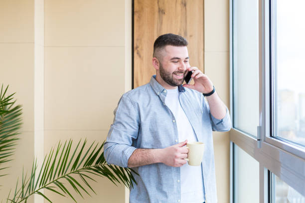 istock Young man at home talking on the phone, drinking coffee by the window, looking at the camera, smiling, quarantined 1368643051