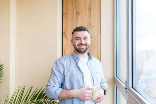 istock Young man at home, drinking coffee by the window, looking at the camera, smiling, quarantined 1368643053