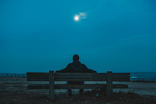 A man sitting in a bench looking at the horizon with the moon on the top