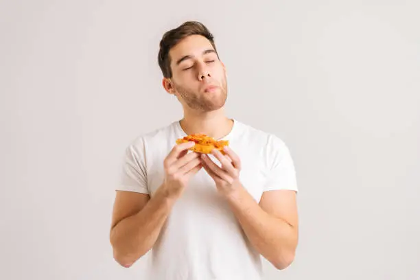 Photo of Portrait of satisfied young man with enjoying eating delicious slice of pizza, with closed eyes from pleasure on white isolated background.