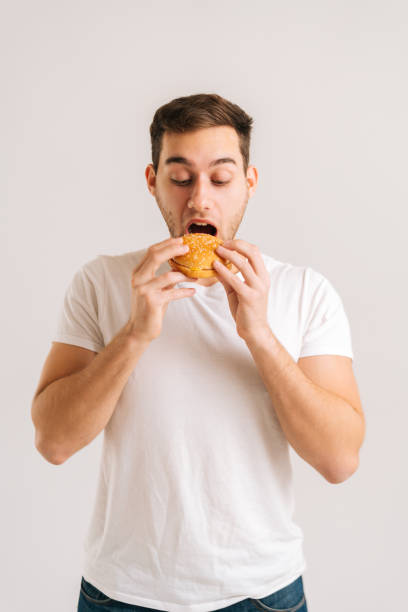 scatto verticale di un bel giovane con il piacere di mangiare delizioso hamburger su sfondo bianco isolato. - burger bun sandwich bread foto e immagini stock