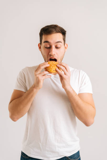 흰색 고립 된 배경에 맛있는 햄버거를 먹는 즐기는 잘 생긴 젊은 남자의 수직 샷. - hamburger bun bread isolated 뉴스 사진 이미지
