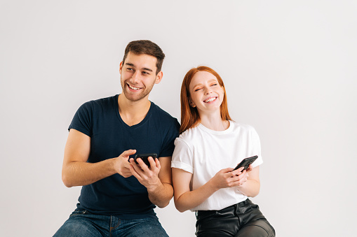 Studio shot of happy young couple laughing using mobile phone, reading good news in social network or email on white isolated background. Communication relationship concept