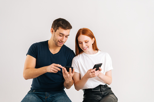 Studio shot of young handsome man showing new mobile software app to smiling girlfriend on white isolated background. Happy black family couple sharing photos or contacts, using smartphones.