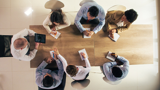 Directly above shot of multiracial group of business people sitting around a conference table, with open notebooks, taking notes and listening to their manager share project data during a corporate meeting.