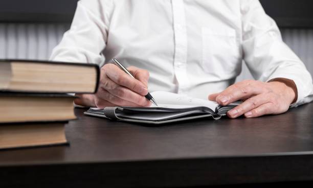 istock Man sitting at office desk with books or codes and taking notes in diary. Man hands closeup. Planning or information analysis concept. 1368649385