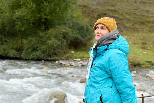 An adult woman in a warm jacket and hat walks along a mountain river and admires nature.