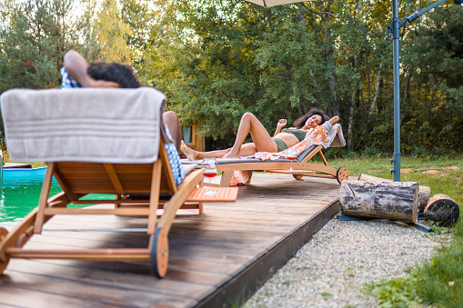 Hispanic man and woman in mid 40s sitting on sun loungers and relaxing by the swimming pool in Boutique Glamping Divja DiVine in Slovenia.