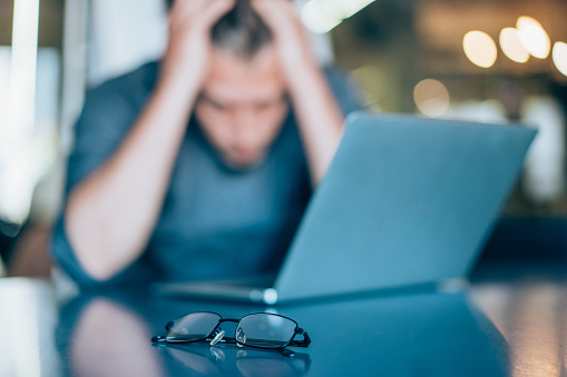 Shot of stressed businessman sitting at his desk and holding head with hands. Overworked businessman sitting in front of laptop and holding head. Focus is on the eyeglasses.