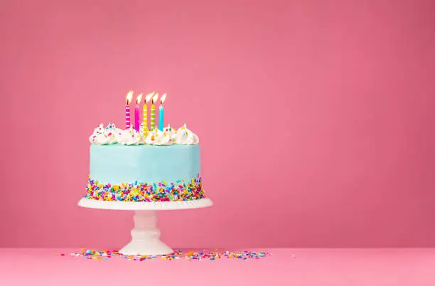 Blue Birthday cake with five colourful Birthday candles and  sprinkles over a pink background.