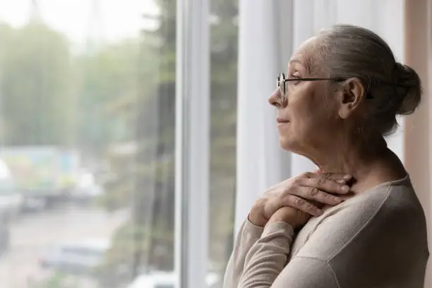 Side view frustrated depressed elderly mature retired woman in eyewear looking in distance out of window, suffering from loneliness or personal problems at home, recollecting memories or mourning.