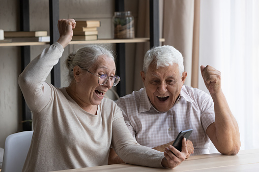 Happy laughing euphoric middle aged old married family couple looking at smartphone screen, reading email or message with amazing news, feeling excited getting online lottery auction win notification.