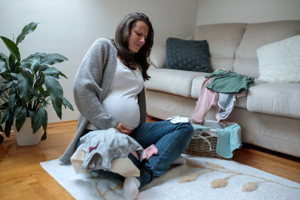 Woman in ninth month of pregnancy experiencing contractions and severe pain sitting in a living room Young Caucasian woman in ninth month of pregnancy experiencing contractions and severe pain while sorting baby clothing in a living room muscular contraction stock pictures, royalty-free photos & images