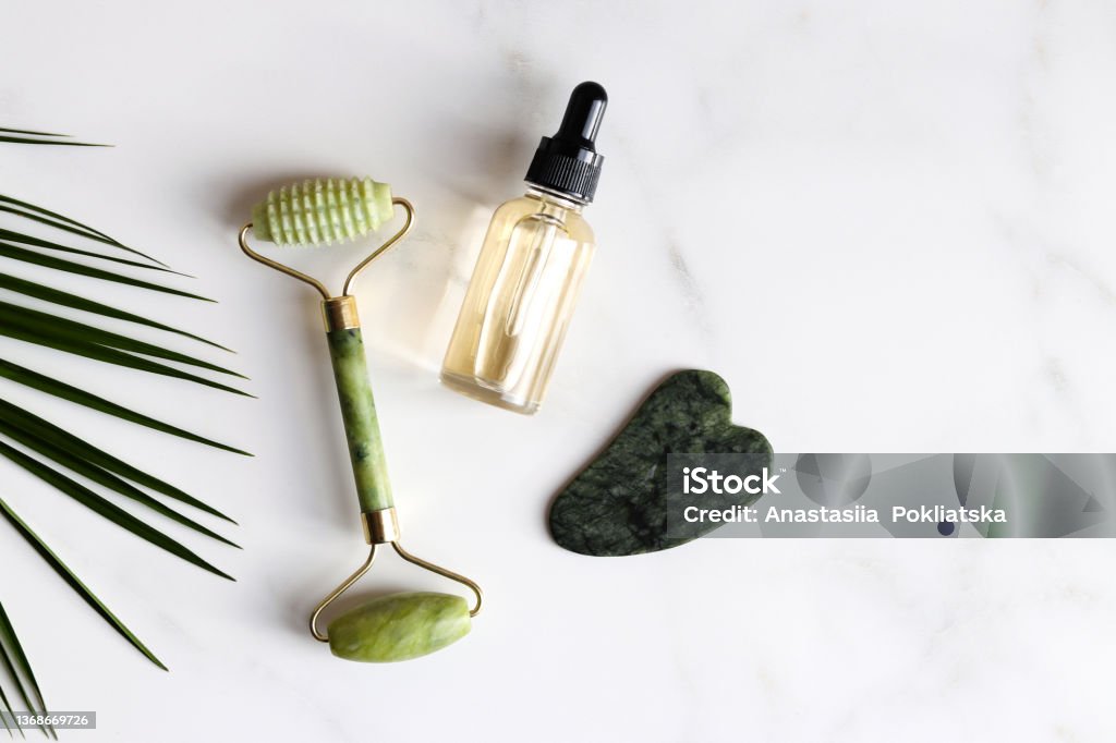 Special devices for Gua Sha. Jade face roller, stone for massage and essential oil on marble table.Traditional Chinese facial massage. Beauty and health care concept. Top view Special devices for Gua Sha. Jade face roller, stone for massage and essential oil on marble table.Traditional Chinese facial massage. Beauty and health care concept. Top view. Anti Aging Stock Photo