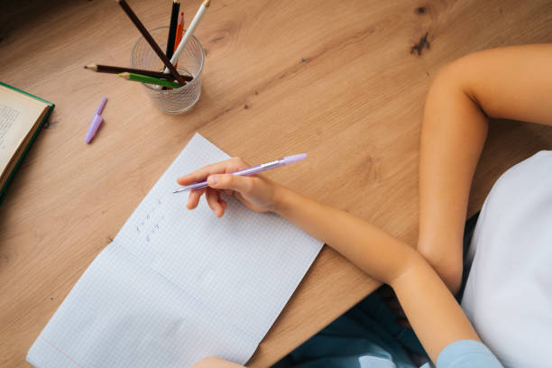 istock Close-up high-angle view of primary daughter doing homework writing in copybook with pen with young mother sitting at home table. 1368698468