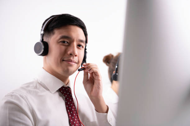 Asian call center operator in customer service department in office. Smiling and friendly Asian call center operator wearing headset with microphone sitting with computer in customer service department in office. Helpful operator dispatcher wearing headset assisting customers in customer support service taking phone calls at office. dispatcher stock pictures, royalty-free photos & images