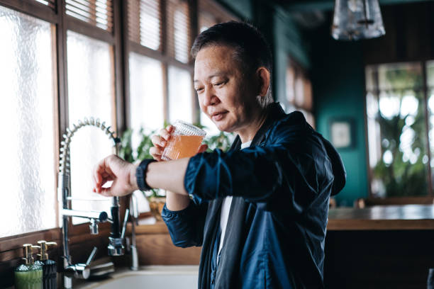 Senior Asian man in sportswear drinking a glass of pink fruit juice over the kitchen counter after exercising at home, checking workout data on fitness tracker app on smartwatch. Staying hydrated while taking a break from daily training hombre asiático mayor en ropa deportiva bebiendo un vaso de jugo de fruta rosa sobre el mostrador de la cocina después de hacer ejercicio en casa, verificando los datos de entrenamiento en la aplicación de seguimiento de ejercicios en el reloj inteligen