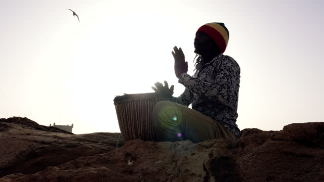 African percussion music, silhouette of a Senegalese man playing a traditional djembe drum with his hands at sunset.