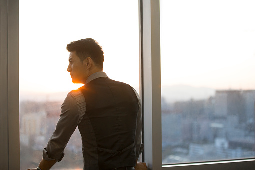Handsome Chinese businessman standing thoughtfully in his office and looking out of the window