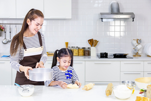 Happy Asian mother teaching her young daughter to bread baking in white modern kitchen while sieving flour for mixing