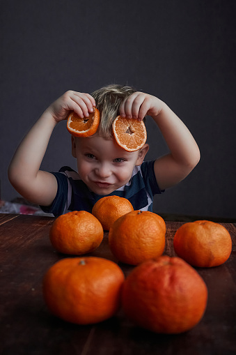 the kid sits at the table on a dark background and is going to eat fresh oranges and indulges