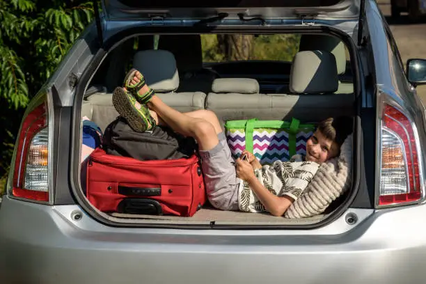 Cute little boy laying on the back of the bags and baggage in the car trunk ready to go on vacation with happy expression. Kid resting playing on smartphone.