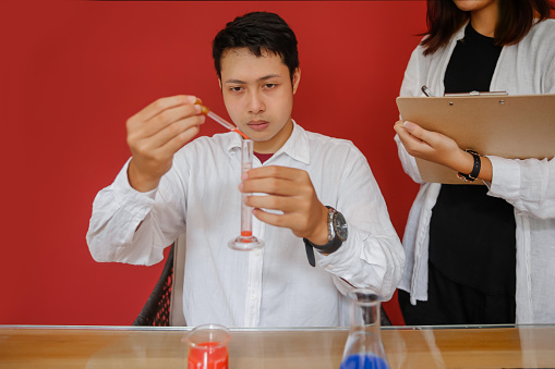 Close up shot of a focus young male scientist pouring a chemical liquid from a pipette into a graduated cylinder ,while his colleague writing notes beside him.