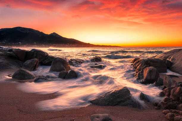 Sun setting over a rocky cove at Aregno Plage in the Balagne region of Corsica with the village of Algajola in the distance