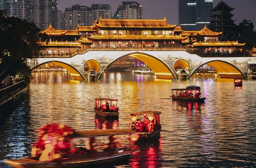 Stream of little tour boats with red lanterns on the Jinjiang River at night, passing the iconic, traditional and illuminating Anshun Bridge in Chengdu, Sichuan, China. Chengdu a vibrant mega city in China.