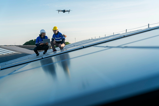 Specialist technician professional engineercontrol drone checking top view of installing solar roof panel on the factory rooftop under sunlight. Engineers holding tablet check solar roof.