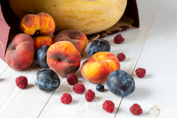 A white bowl filled with assorted fruits including plums, raspberries, and pears sits on a wooden table. A freshly cut peach rests on a wooden cutting board nearby, inviting a soothing autumn vibe. Copy space for text, table top view - food and drink, fruit salad