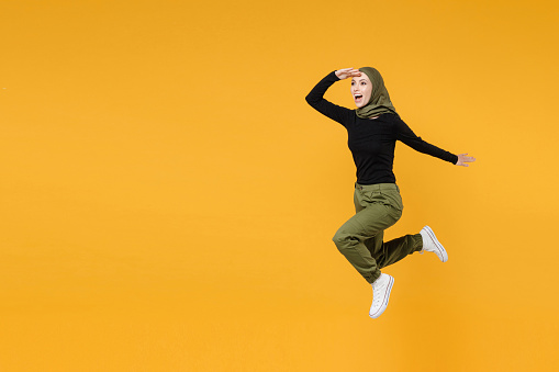 Full length of excited young arabian muslim woman wearing hijab black green clothes jumping looking far away distance isolated on yellow background studio portrait. People religious lifestyle concept