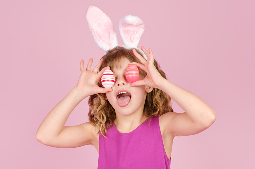 Closeup portrait of cheerful little girl in Easter bunny ears smiling while covering eyes with colorful eggs against pink background