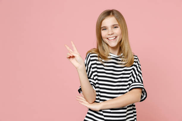 piccola bionda sorridente carino bambino bella ragazza 12-13 anni che indossa una t-shirt oversize a righe mostra vittoria v-sign gesto isolato su sfondo rosa pastello bambini studio . concetto di stile di vita infantile. - teenager 14 15 years 13 14 years cheerful foto e immagini stock