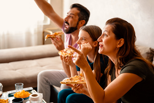Family watching game on TV and eating pizza. Parents and daughter watching Television at home. Family watching sport matches enjoying fast food