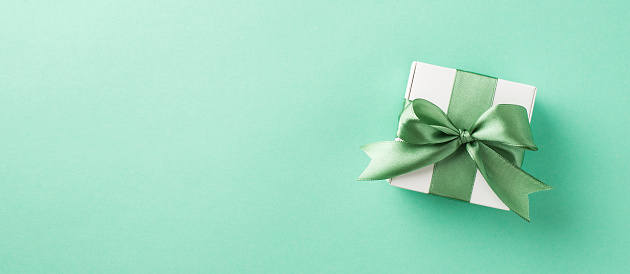 Top view photo of a small white gift box with cute silk green bow on the pastel turquoise isolated background copyspace