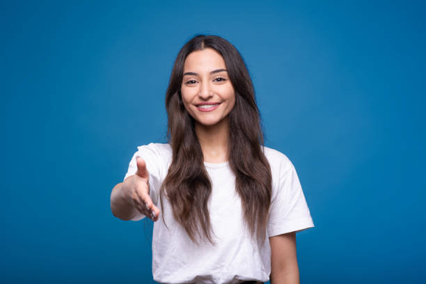 garota morena branca ou árabe atraente em uma camiseta branca oferecendo um aperto de mão para parceria isolada em um fundo de estúdio azul. - isolated smiling business person handshake - fotografias e filmes do acervo