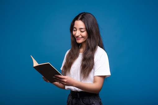 Attractive caucasian or arab brunette girl in a white t-shirt reading an interesting book and smiling isolated on a blue studio background.