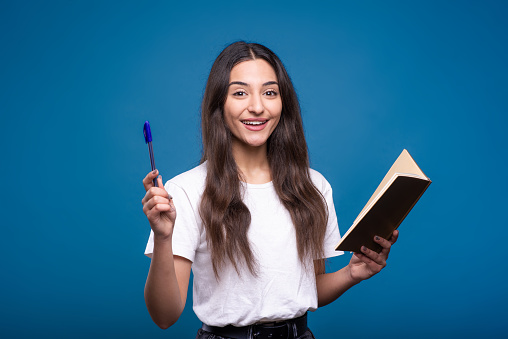 Attractive caucasian or arab brunette girl in a white t-shirt is shocked holding a notepad in her hand and writing, she has an idea isolated on a blue studio background.