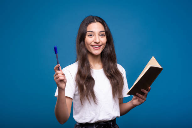 Attractive caucasian or arab brunette girl in a white t-shirt is shocked holding a notepad in her hand and writing, she has an idea isolated on a blue studio background. Attractive caucasian or arab brunette girl in a white t-shirt is shocked holding a notepad in her hand and writing, she has an idea isolated on a blue studio background. blue pen stock pictures, royalty-free photos & images