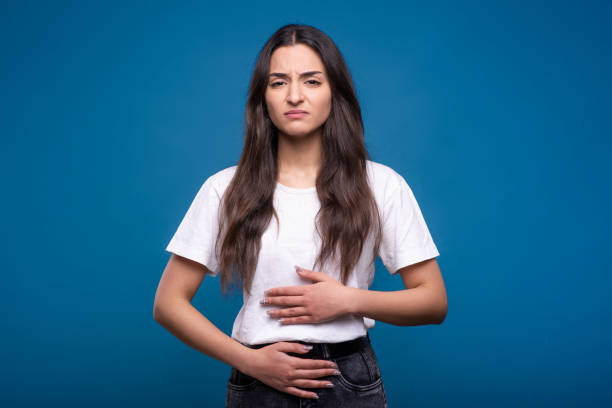 joven y atractiva chica morena caucásica o árabe con camiseta blanca está insatisfecha debido al dolor de estómago, dolor de estómago aislado sobre el fondo azul del estudio. - holding belly fotografías e imágenes de stock