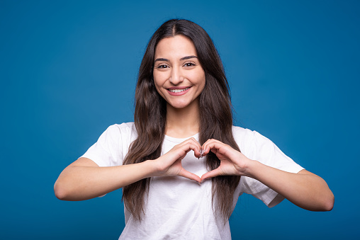 Young and attractive caucasian or arab brunette girl in white t-shirt showing love gesture near heart - valentine shape isolated on blue studio background.
