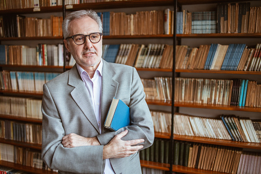 University teacher posing for a portrait in an university library with a book under his arm, wearing eyeglasses.