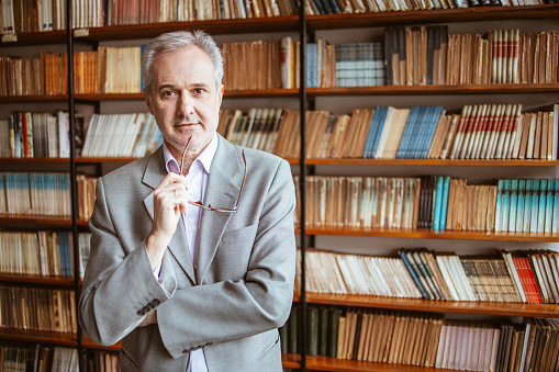 University teacher posing for a portrait in an university library holding an eyeglasses.