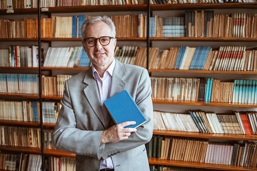 University teacher posing for a portrait in an university library holding a book in his arm, wearing eyeglasses.