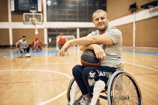 Happy disabled athlete in wheelchair holding basketball on sports court and looking at camera.
