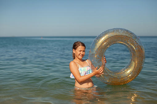 A tanned smiling girl stands almost waist-deep in the azure water of the ocean and holds a inflatable swimming ring on a sunny day in summer, space for text.